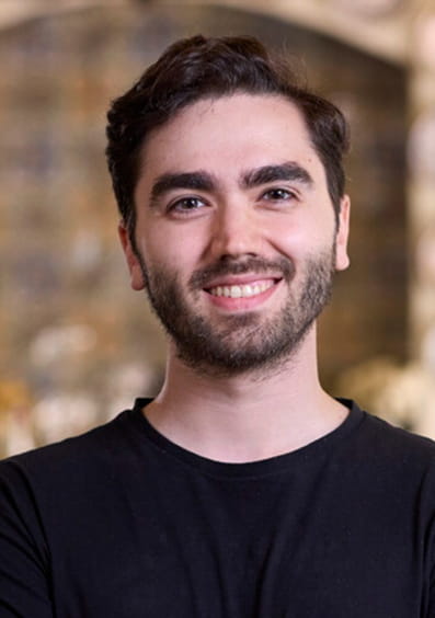 Smiling man with dark hair and beard wearing a black shirt standing indoors.