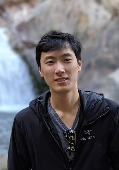 Young man with black hair wearing a dark jacket and sunglasses standing near a waterfall.