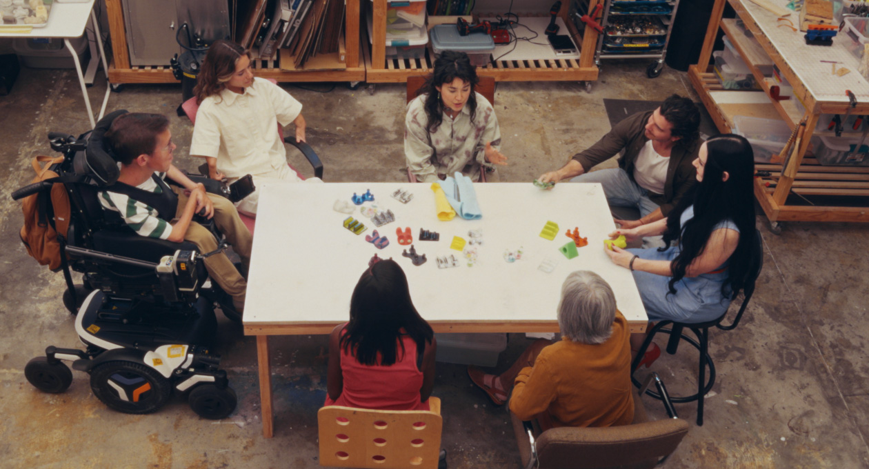 Um grupo de pessoas com diferentes necessidades de mobilidade sentadas juntas a uma mesa, enquanto observam uma variedade de acessórios coloridos adaptáveis para smartphones